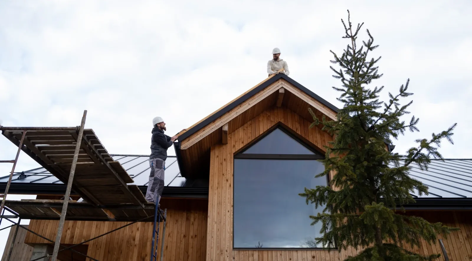 two workers wearing hats installing the roof of a wooden house, one standing on scaffolding and the other on the roof peak